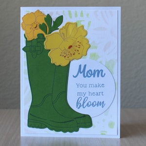 Puede incluir: Tarjeta de felicitación hecha a mano con botas de agua verdes y flores amarillas. La tarjeta incluye el texto "Mom, You make my heart bloom" en escritura azul sobre un óvalo blanco.