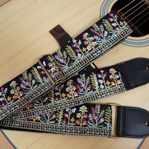 May include: Black guitar strap with colorful floral embroidery. The strap features a repeating pattern of flowers and foliage in shades of yellow, pink, and green. The strap has black leather ends and brass-colored hardware.