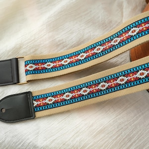 Könnte beinhalten: Ein beigefarbener Gitarrengurt mit einem farbenfrohen geometrischen Muster in Rot, Blau und Weiß. Der Gurt hat schwarze Lederenden und goldfarbene Metallbeschläge. Das Design ist auf dem Gurt zentriert.