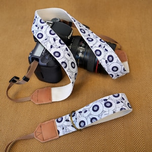 May include: A camera strap featuring a white fabric base with a repeating dark blue pattern of circular parasols. The strap includes tan leather end tabs, brown nylon webbing connectors, and a metal ring, attached to a black digital camera body.