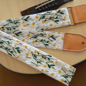 May include: Cream-colored guitar strap with embroidered white daisies and yellow centers, accented by green leaves. The strap features tan leather ends and is draped over a wooden acoustic guitar.
