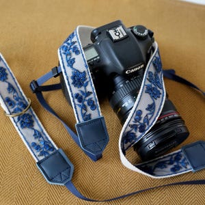 Könnte beinhalten: Eine schwarze Canon DSLR-Kamera mit einem 24-105mm Objektiv und einem dekorativen Kameragurt. Der Gurt besteht aus hellbeigem Stoff mit einer dunkelblauen floralen Spitzenauflage, dunkelblauen Lederenden und einem dunkelblauen Nylonband.