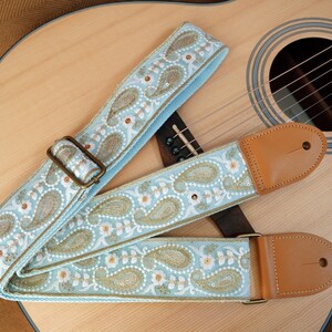 May include: A light blue guitar strap with a paisley pattern in white, gold, and brown. The strap has brown leather ends and a bronze-colored metal buckle. The strap is lying on a wooden acoustic guitar.
