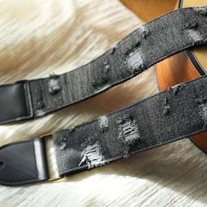 Könnte beinhalten: Nahaufnahme eines Gitarrengurts aus Used-Denim. Der Gurt ist schwarz mit absichtlichen Rissen und Löchern und hat schwarze Lederenden mit einem Loch zur Befestigung. Eine goldfarbene Metallschnalle ist sichtbar. Der Hintergrund ist eine hellfarbige, strukturierte Oberfläche.