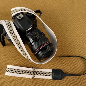 May include: A black Canon DSLR camera with a zoom lens attached to a cream-colored fabric camera strap. The strap features a decorative woven pattern in metallic gold and brown thread, with black leather accents and a brass-colored metal ring.