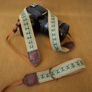 May include: A camera strap featuring a beige woven base with a repeating light green floral pattern, accented by dark brown leather end tabs and thin brown nylon webbing straps, shown attached to a black DSLR camera with a lens.