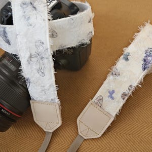May include: A white camera strap with a butterfly pattern. The strap has a textured, fringed edge and is paired with a black camera lens. The butterfly design features blue and purple accents. The strap is attached to the camera with a light beige leather tab.