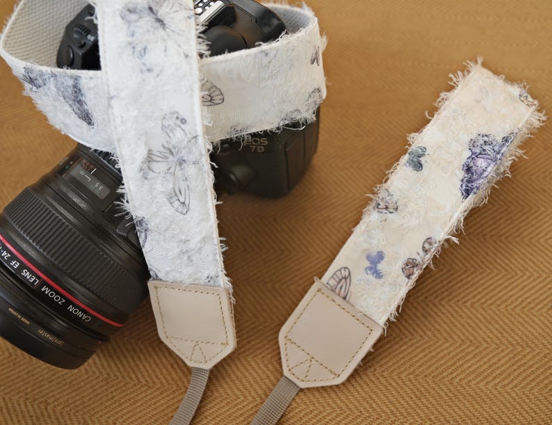 May include: A white camera strap with a butterfly pattern. The strap has a textured, fringed edge and is paired with a black camera lens. The butterfly design features blue and purple accents. The strap is attached to the camera with a light beige leather tab.