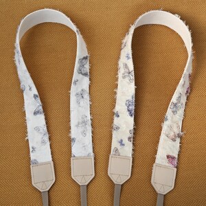 May include: Two camera straps with a white background and a butterfly pattern in shades of blue and purple. The straps have a textured, fringed edge and beige leather ends with gray connectors. The straps are displayed on a golden-brown surface.
