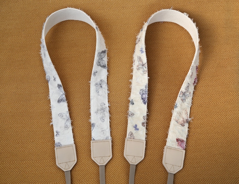 May include: Two camera straps with a white background and a butterfly pattern in shades of blue and purple. The straps have a textured, fringed edge and beige leather ends with gray connectors. The straps are displayed on a golden-brown surface.
