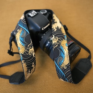 May include: A black camera strap featuring a repeating pattern of stylized blue and gold ocean waves. The strap is attached to a black Canon DSLR camera with a zoom lens, showcasing the detailed graphic print across the fabric surface.