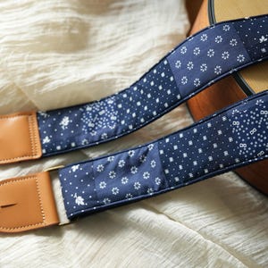 Könnte beinhalten: Zwei marineblaue Gitarrengurte mit einem Patchwork-Design mit weißen floralen und geometrischen Mustern. Die Gurte haben braune Lederenden und goldfarbene Metallbeschläge.