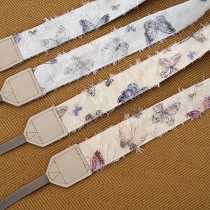 May include: Four camera straps with butterfly patterns. The straps have a cream-colored base with butterfly illustrations in shades of gray, purple, and pink. Each strap has a light gray nylon strap and a beige leather connector.