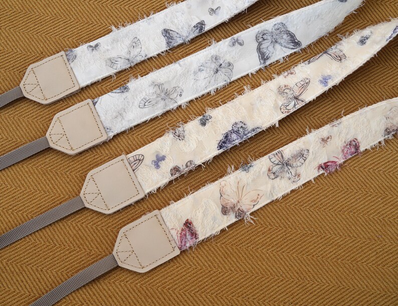 May include: Four camera straps with butterfly patterns. The straps have a cream-colored base with butterfly illustrations in shades of gray, purple, and pink. Each strap has a light gray nylon strap and a beige leather connector.