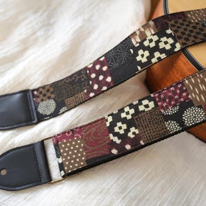 Könnte beinhalten: Ein Gitarrengurt mit Patchwork-Design in Braun-, Schwarz- und Burgundertönen. Der Gurt hat schwarze Lederenden und eine Metallschnalle. Das Design umfasst florale und geometrische Muster.