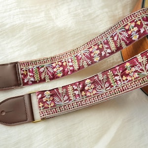 Könnte beinhalten: Burgunderfarbener Gitarrengurt mit aufwendiger Blumenstickerei in Gelb, Grün und Rosa. Braune Lederenden und goldfarbene Hardware. Das verzierte Design ist für Akustik- oder E-Gitarren geeignet.