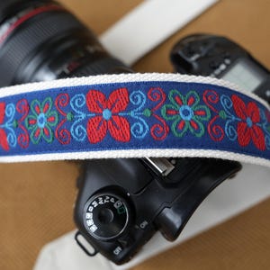 Könnte beinhalten: Ein Kameragurt mit einem blauen Stoffband, das ein gesticktes Blumenmuster in Rot, Hellblau und Grün aufweist und von weißen Webkanten gesäumt ist. Der Gurt liegt über einer schwarzen Digitalkamera mit Objektiv und sichtbarem Einstellrad.