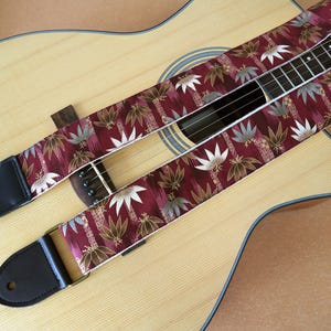 May include: Acoustic guitar with a burgundy strap featuring a floral pattern in white, gold, and gray. The strap has black leather ends and gold-colored metal hardware. The guitar is light brown.