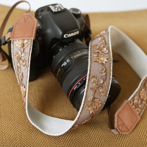May include: A black Canon DSLR camera with a 24-105mm lens attached, featuring a decorative camera strap. The strap is made of a sheer, taupe-colored mesh fabric with intricate gold floral embroidery, tan leather end tabs, and a white backing.