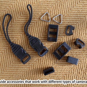 May include: A collection of black camera accessories, including buckles, straps, and metal triangle rings, are arranged on a textured tan surface. The image includes the text: "We provide accessories that work with different types of camera eyelets."