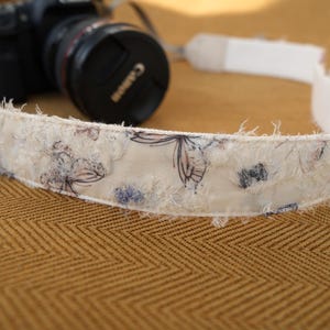 May include: A cream-colored camera strap with a floral pattern and frayed edges. The strap is decorated with blue and purple floral designs. A black Canon camera is visible in the background.