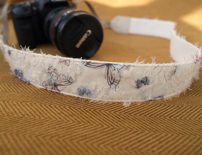 May include: A cream-colored camera strap with a floral pattern and frayed edges. The strap is decorated with blue and purple floral designs. A black Canon camera is visible in the background.