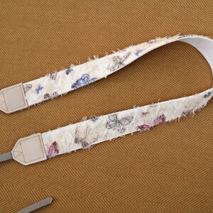 May include: A cream-colored camera strap with a butterfly pattern in shades of purple, blue, and pink. The strap has a textured, fringed edge and light gray leather connectors.