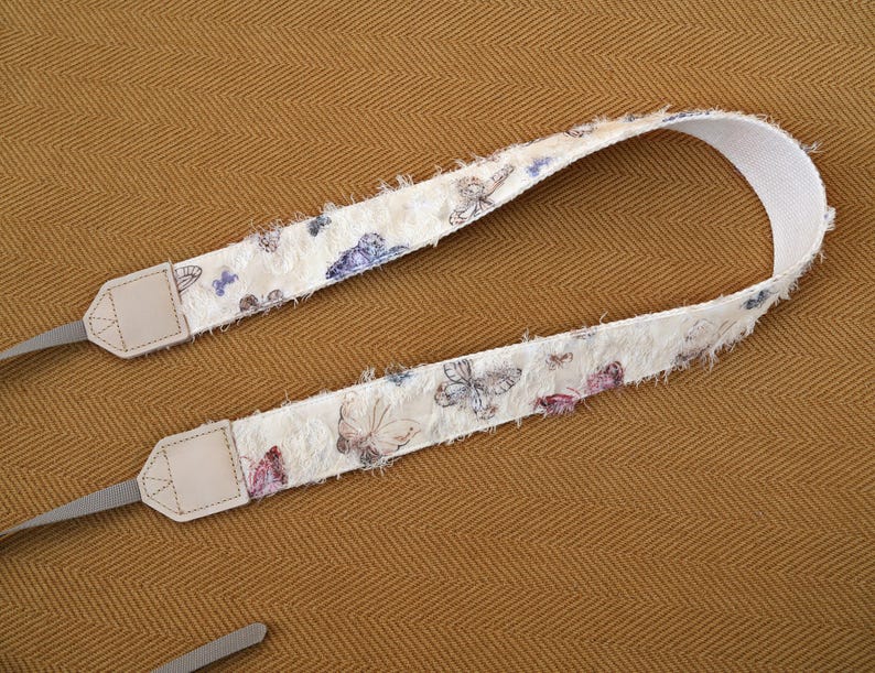 May include: A cream-colored camera strap with a butterfly pattern in shades of purple, blue, and pink. The strap has a textured, fringed edge and light gray leather connectors.
