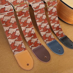 Könnte beinhalten: Vier Gitarrengurte mit einem sich wiederholenden Tessellationsmuster aus orangefarbenen Vögeln und hellbeigen Fischen. Jeder Gurt verfügt über verstellbare Metallbeschläge und ein Lederende in Hellbraun, Dunkelbraun, Blau oder Schwarz.