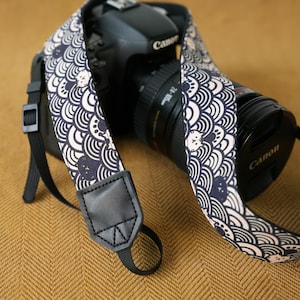 May include: A camera strap featuring a repeating pattern of dark blue and white stylized waves interspersed with small cat faces. The strap includes black faux leather end tabs with black nylon webbing connectors attached to a black Canon camera.