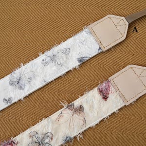 May include: Two camera straps with butterfly patterns. One strap features a white background with gray butterflies, the other has a cream background with pink and purple butterflies. Both straps have tan leather ends and brown nylon straps.