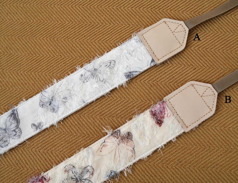 May include: Two camera straps with butterfly patterns. One strap features a white background with gray butterflies, the other has a cream background with pink and purple butterflies. Both straps have tan leather ends and brown nylon straps.