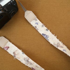 May include: Two camera straps with a cream-colored, butterfly-patterned fabric. The straps have frayed edges and tan leather ends. A black camera is visible in the upper left corner, resting on a textured, golden-brown surface.