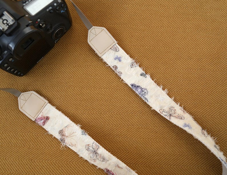 May include: Two camera straps with a cream-colored, butterfly-patterned fabric. The straps have frayed edges and tan leather ends. A black camera is visible in the upper left corner, resting on a textured, golden-brown surface.