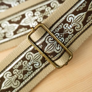 May include: Close-up of a beige and brown guitar strap with ornate embroidered detailing. The strap features a repeating pattern of stylized floral designs in brown and cream, with a brass-colored metal buckle.