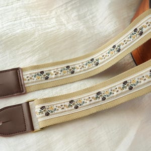 Könnte beinhalten: Zwei beige Gitarrengurte mit floraler Stickerei. Die Gurte haben braune Lederenden und goldfarbene Metallbeschläge. Das Blumenmuster enthält gelbe, braune und grüne Akzente auf einem weißen Streifen.