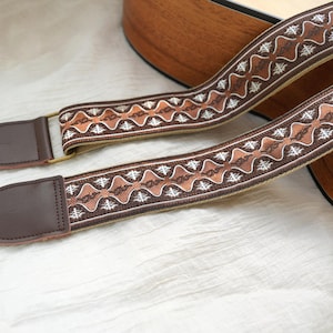 May include: A brown and tan guitar strap with a decorative woven pattern. The strap features brown leather ends and metal hardware for attachment. The design includes repeating geometric shapes in shades of brown, tan, and white.