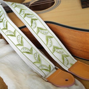 May include: White guitar strap with green leaf and pearl-like accents. The strap has brown leather ends and silver hardware. The strap is resting on a light brown acoustic guitar.