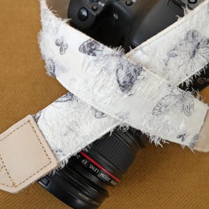 May include: A white camera strap with a butterfly pattern in gray and black. The strap has a frayed edge and a light tan leather end piece. The strap is wrapped around a black camera lens.