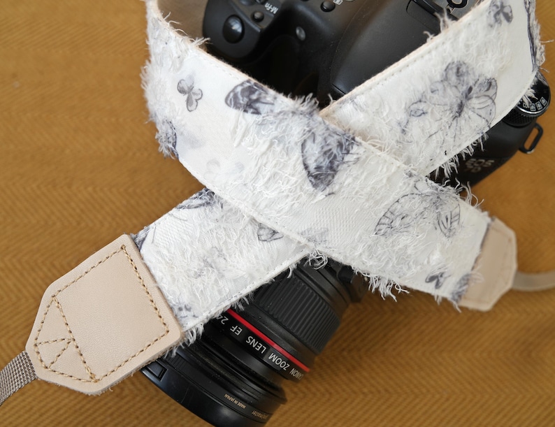 May include: A white camera strap with a butterfly pattern in gray and black. The strap has a frayed edge and a light tan leather end piece. The strap is wrapped around a black camera lens.
