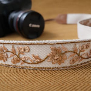 May include: Cream-colored camera strap with embroidered gold floral design. The strap is made of a woven material and features a repeating pattern of flowers and leaves. The strap is shown with a black camera and lens.