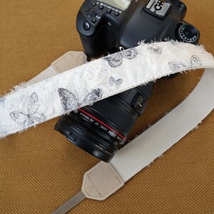 May include: A white camera strap with a butterfly pattern in gray and black is draped over a black DSLR camera. The strap has a frayed edge and is made of a soft, textured material. The camera lens is black with red lettering.