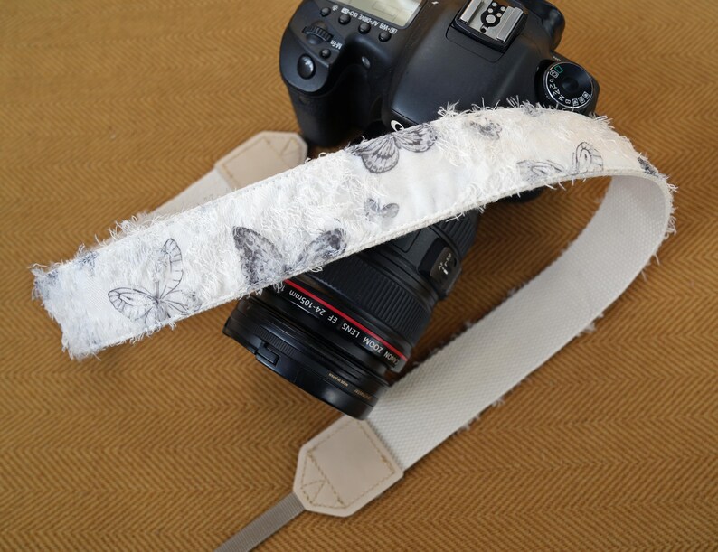 May include: A white camera strap with a butterfly pattern in gray and black is draped over a black DSLR camera. The strap has a frayed edge and is made of a soft, textured material. The camera lens is black with red lettering.