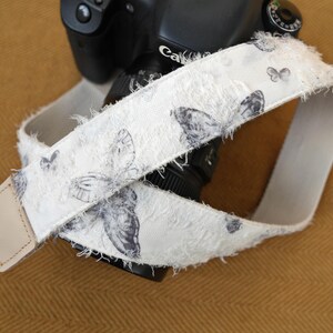 May include: A white camera strap with a butterfly pattern. The strap is made of a textured fabric with frayed edges. The butterflies are in shades of gray. The strap is attached to a black camera.