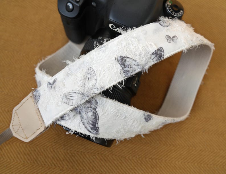 May include: A white camera strap with a butterfly pattern. The strap is made of a textured fabric with frayed edges. The butterflies are in shades of gray. The strap is attached to a black camera.