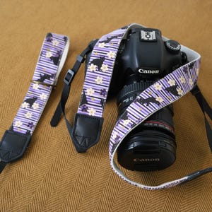 Könnte beinhalten: Eine schwarze Canon DSLR-Kamera mit einem 24-105mm Objektiv und einem Stoff-Kameragurt mit violett-weißem Streifenmuster, schwarzen Katzensilhouetten und weißen Blüten. Ein passender, abgenommener Kameragurt liegt daneben.