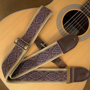 May include: A guitar strap featuring a beige woven base with a repeating purple and blue diamond-shaped pattern. The strap includes dark brown leather end tabs with circular holes and an adjustable antique brass metal slider for length adjustment.