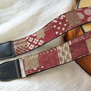 Op de afbeelding: Een gitaarband met een patchwork ontwerp in bordeauxrood, beige en wit. De band heeft zwarte leren uiteinden en een messingkleurige gesp. De stof heeft bloemen- en geometrische patronen.