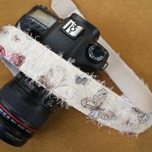 May include: A black DSLR camera with a cream-colored strap featuring a butterfly pattern. The strap has a frayed edge and is draped over the camera body and lens. The lens is black with red lettering.