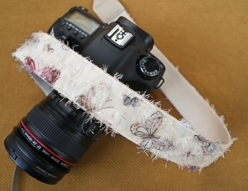 May include: A black DSLR camera with a cream-colored strap featuring a butterfly pattern. The strap has a frayed edge and is draped over the camera body and lens. The lens is black with red lettering.